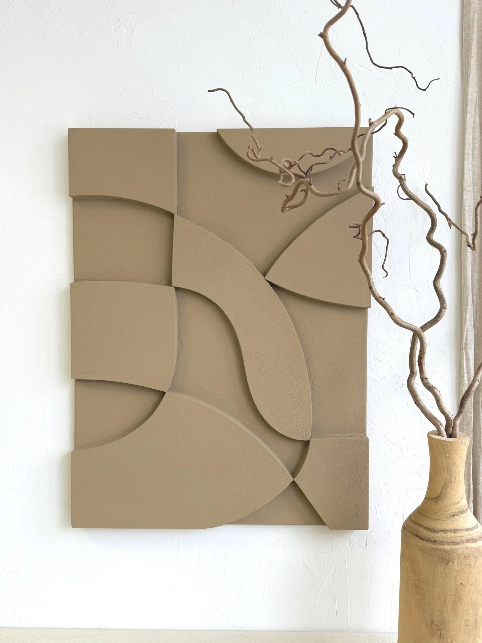 Beige abstract wall art piece on a white wall with a vase and branches.